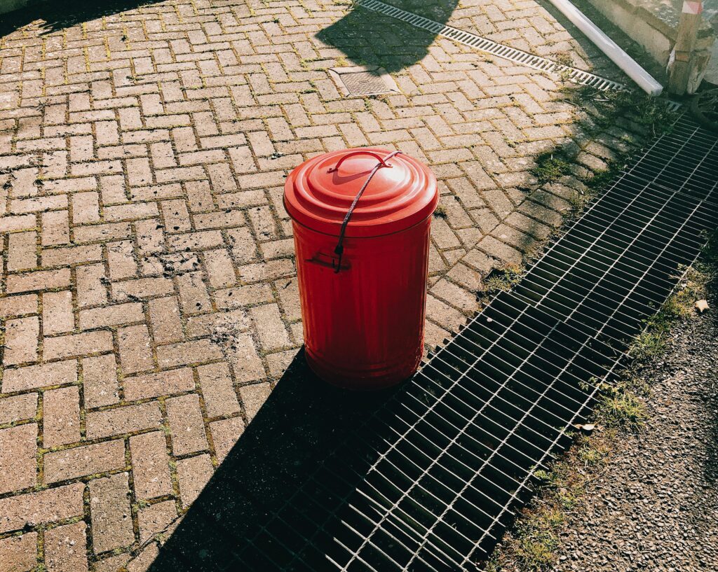 High angle of red metal trash can placed on pavement near sewer grates in city street in sunny summer day